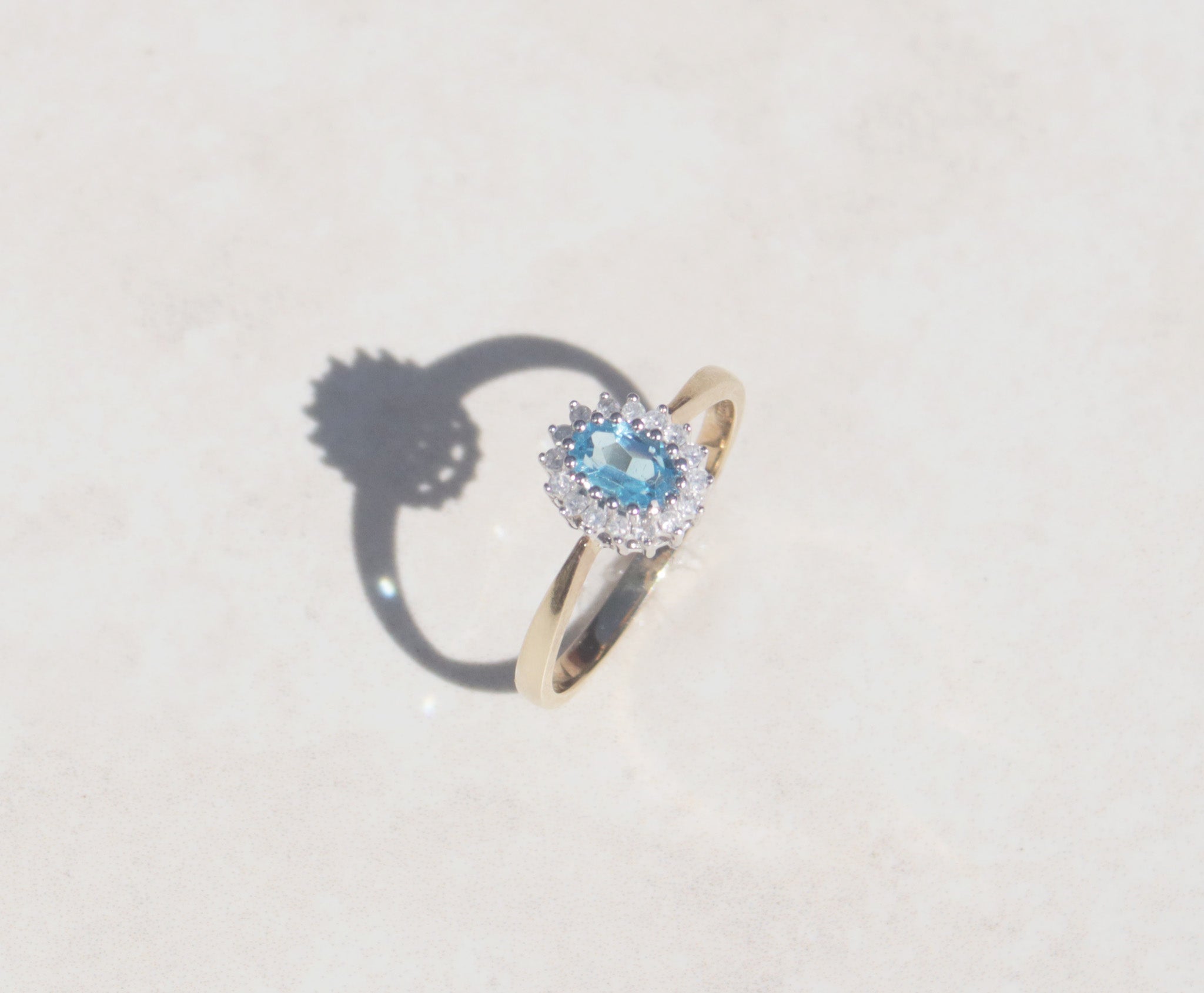 This classic 9ct yellow gold ring features a blue topaz silhouetted by a cluster of round brilliant cut diamonds on a dainty yellow gold band. The oval cut blue topaz has a captivating fire and strong Swiss blue colour accentuated by the open gallery and 9ct white gold claw setting. The claw set diamond cluster features a family of sixteen round brilliant cut diamonds, with a total carat weight of 0.32 ct.