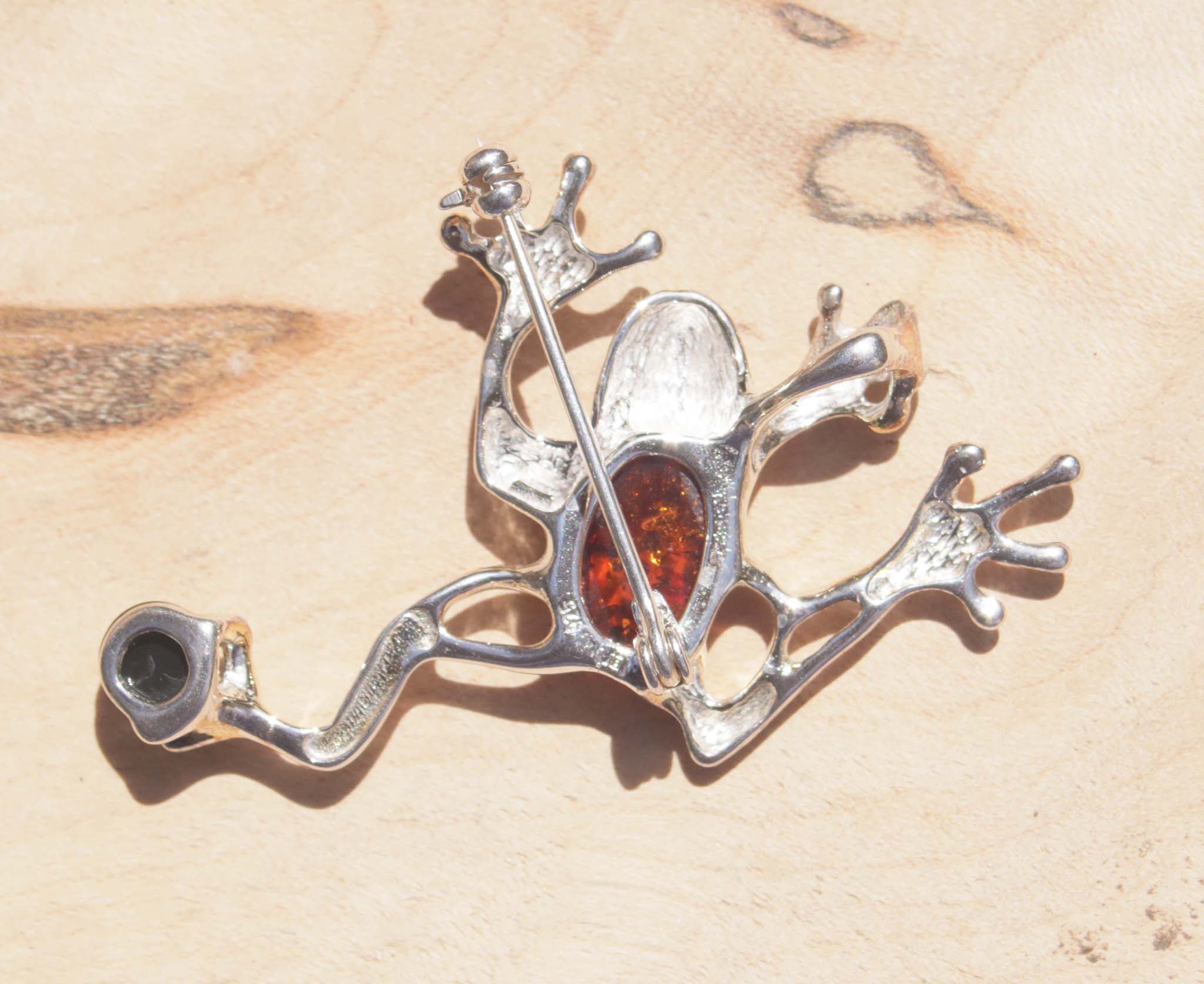 A sterling silver cognac and green amber jumping frog brooch. This brooch features a variety of natural amber, a material made from the resin of trees. The amber chosen for the body of the frog displays natural inclusions which cleverly reflect the pattern of frogs.