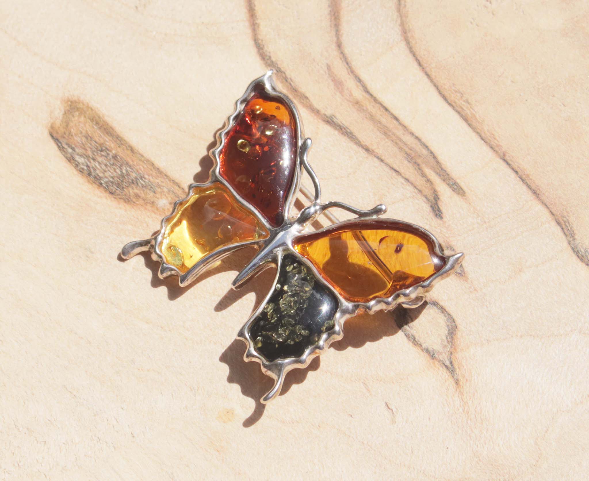 An eye-catching sterling silver amber butterfly brooch. This brooch contains a variety of natural amber, a natural material made from the resin of trees. Set in sterling silver, the amber wings include cognac, honey, orange and green amber.
