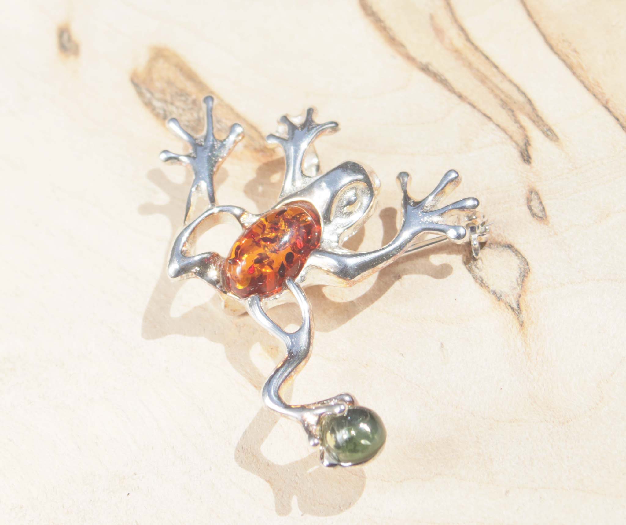 A sterling silver cognac and green amber jumping frog brooch. This brooch features a variety of natural amber, a material made from the resin of trees. The amber chosen for the body of the frog displays natural inclusions which cleverly reflect the pattern of frogs.