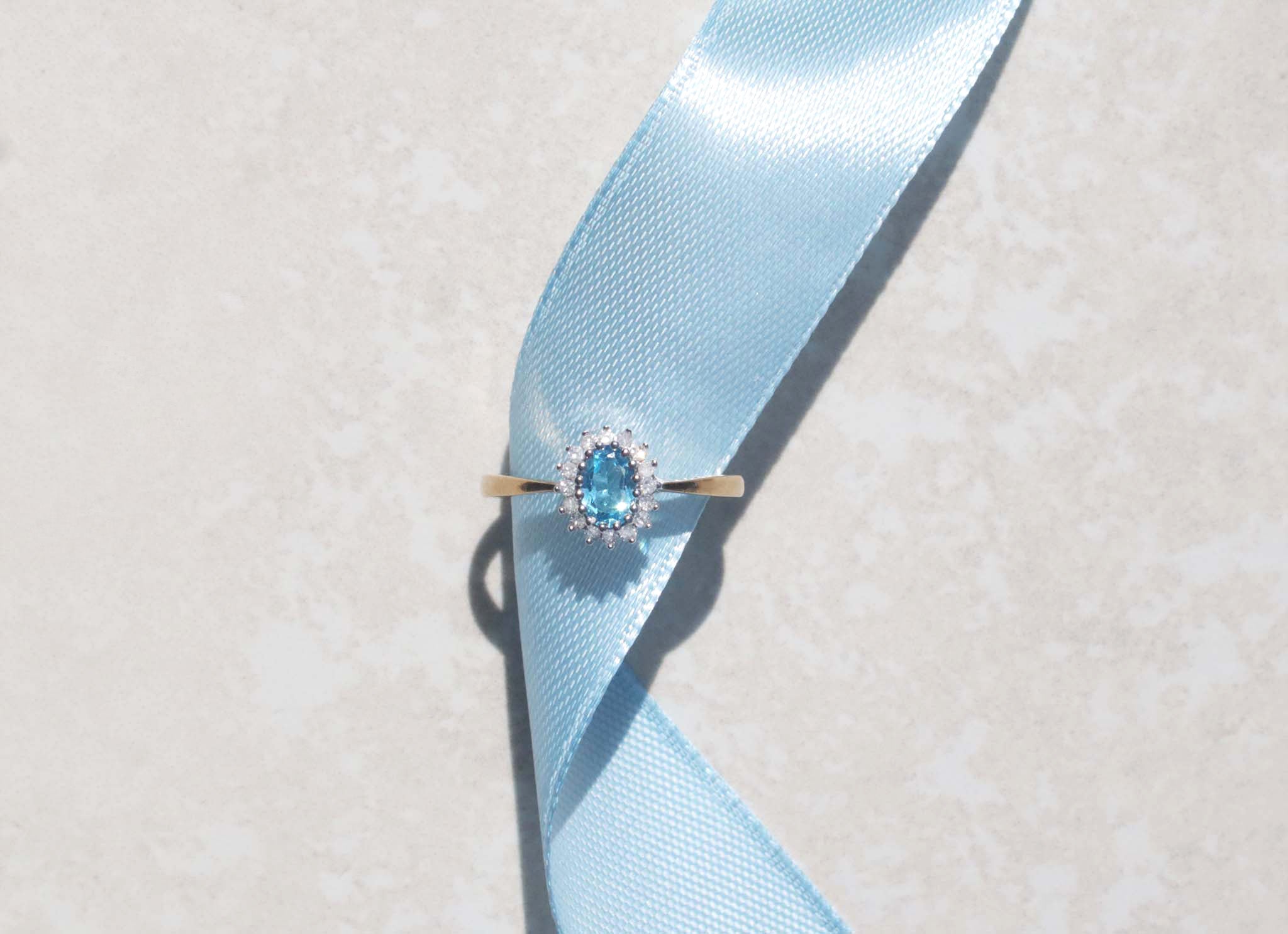 This classic 9ct yellow gold ring features a blue topaz silhouetted by a cluster of round brilliant cut diamonds on a dainty yellow gold band. The oval cut blue topaz has a captivating fire and strong Swiss blue colour accentuated by the open gallery and 9ct white gold claw setting. The claw set diamond cluster features a family of sixteen round brilliant cut diamonds, with a total carat weight of 0.32 ct.
