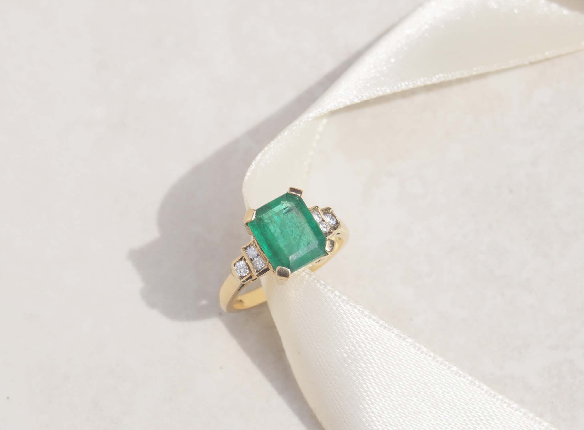The image displays a 18ct yellow gold Art Deco ring features emerald and diamonds.  The rich green emerald measures 3.0ct and takes centre stage among six round brilliant cut diamonds.  18ct yellow gold Gemstone : Emerald  Emerald cut, 3.0cts  Six round brilliant cut diamonds, SI1, F, total carat weight 0.12cts, Weight 5.58g
