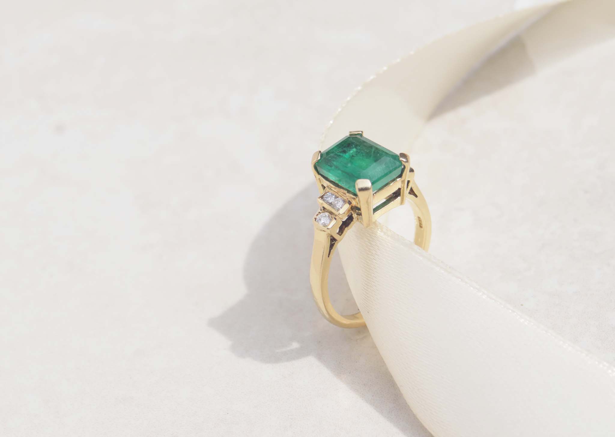 The image displays a 18ct yellow gold Art Deco ring features emerald and diamonds.  The rich green emerald measures 3.0ct and takes centre stage among six round brilliant cut diamonds.  18ct yellow gold Gemstone : Emerald  Emerald cut, 3.0cts  Six round brilliant cut diamonds, SI1, F, total carat weight 0.12cts, Weight 5.58g