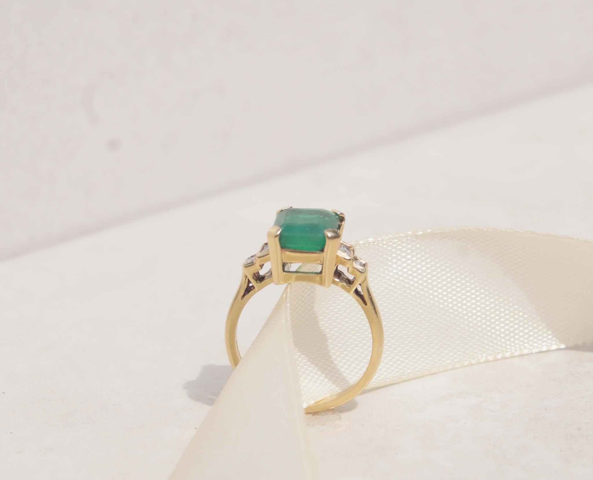 The image displays a 18ct yellow gold Art Deco ring features emerald and diamonds. The rich green emerald measures 3.0ct and takes centre stage among six round brilliant cut diamonds. 18ct yellow gold Gemstone : Emerald Emerald cut, 3.0cts Six round brilliant cut diamonds, SI1, F, total carat weight 0.12cts, Weight 5.58g