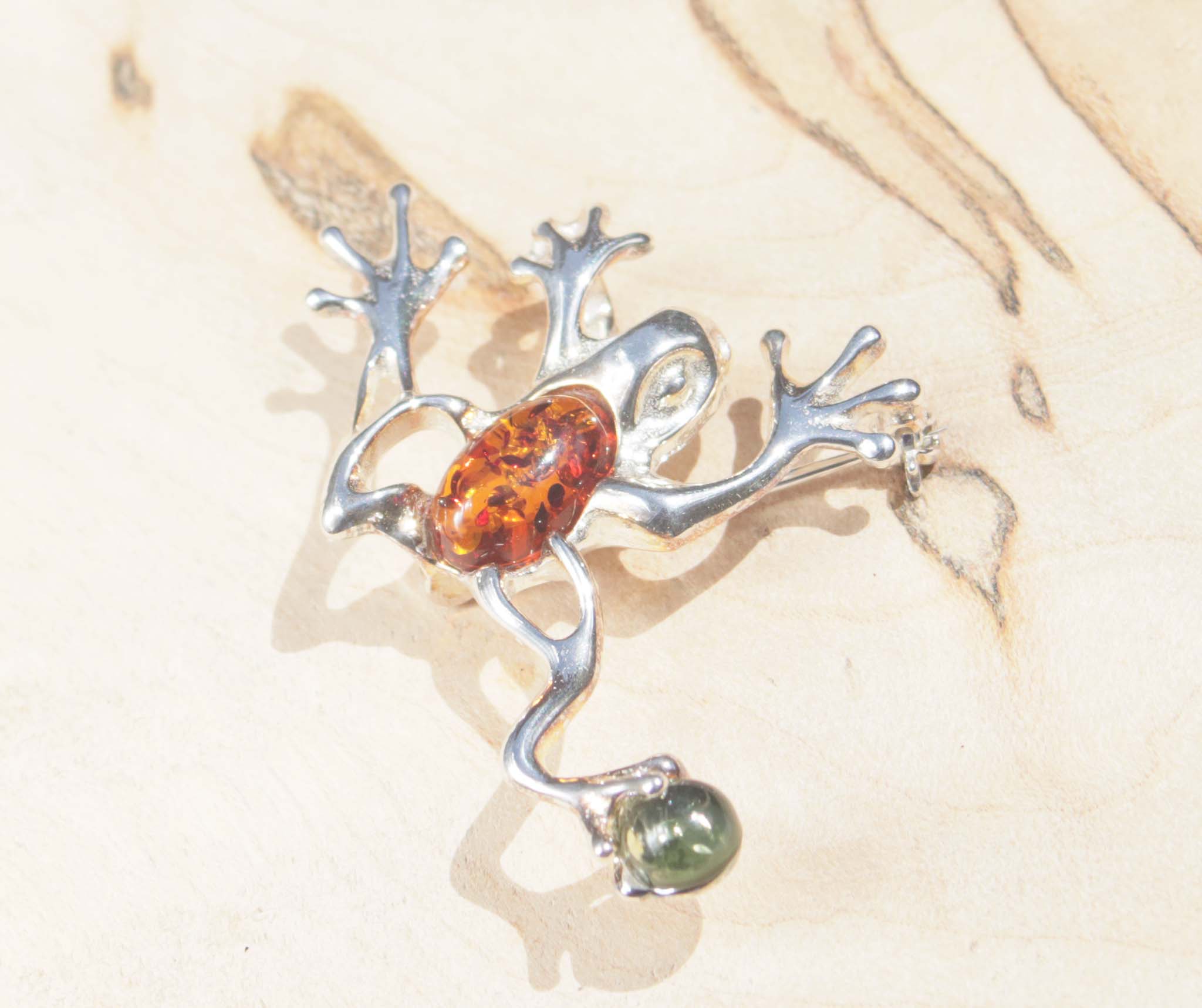 A sterling silver cognac and green amber jumping frog brooch.   This brooch features a variety of natural amber, a material made from the resin of trees. The amber chosen for the body of the frog displays natural inclusions which cleverly reflect the pattern of frogs. 
