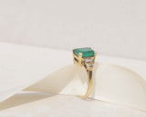The image displays a 18ct yellow gold Art Deco ring features emerald and diamonds.  The rich green emerald measures 3.0ct and takes centre stage among six round brilliant cut diamonds.  18ct yellow gold Gemstone : Emerald  Emerald cut, 3.0cts  Six round brilliant cut diamonds, SI1, F, total carat weight 0.12cts, Weight 5.58g 